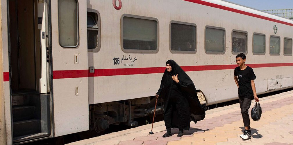 مسافرون يسيرون على رصيف محطة قطار بجانب عربة قطار ذات لون أبيض وشريط أحمر