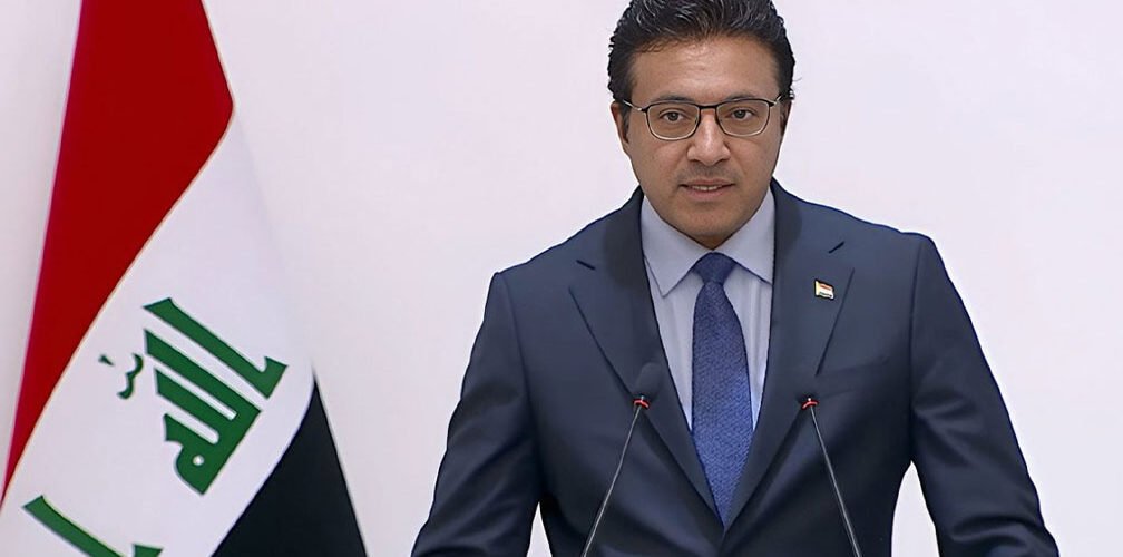 المتحدث باسم الحكومة العراقية باسم العوادي يتحدث خلف الميكروفون وإلى جانبه علم العراق