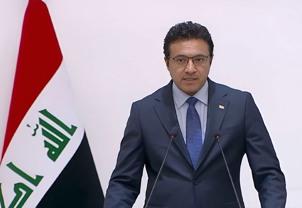 المتحدث باسم الحكومة العراقية باسم العوادي يتحدث خلف الميكروفون وإلى جانبه علم العراق